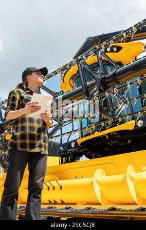Händler oder Lieferant von Landmaschinen. Weiße Frau im karierten Hemd steht neben dem Mähdrescher und verwendet ein digitales Tablet. Stockfoto