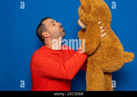 Bärtiger hispanischer Mann in seinen 40ern, der einen roten Pullover trägt, einen Teddybären hochhält, während er Küsse bläst und ihn ankuschelt, isoliert im blauen Studio Stockfoto