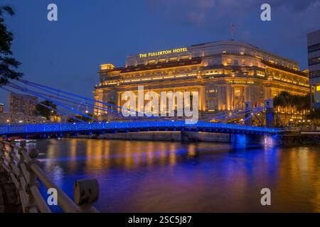 Die tiefblaue Spanne der Cavanagh Bridge verläuft über dem Singapore River unter einem leuchtenden Fullerton Hotel, mit dem Singapore Flyer im Hintergrund Stockfoto