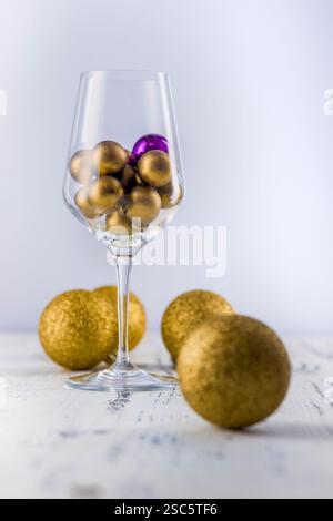 Weinglas gefüllt mit goldenen Kugeln und einer violetten Kugel. Modernes, minimalistisches Stillleben auf einer weiß strukturierten Oberfläche. Skurril und konzeptuell. Perfec Stockfoto