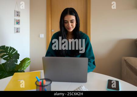 Junge Frau, die von zu Hause aus auf einem Laptop am Tisch arbeitet und lächelt Stockfoto