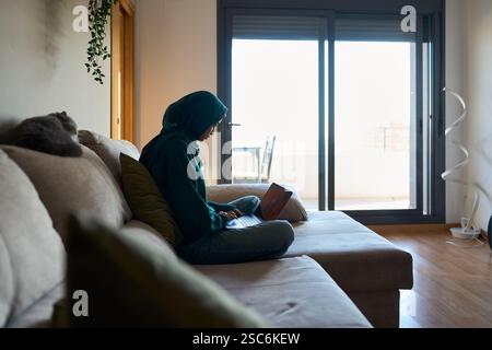 Muslimische Frau, die von zu Hause aus auf einem Laptop arbeitet, mit einer Katze, die neben ihr schläft Stockfoto