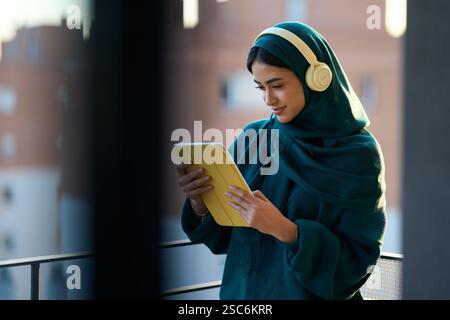 Junge muslimische Frau, die Kopfhörer mit einem digitalen Tablet auf dem Balkon trägt Stockfoto