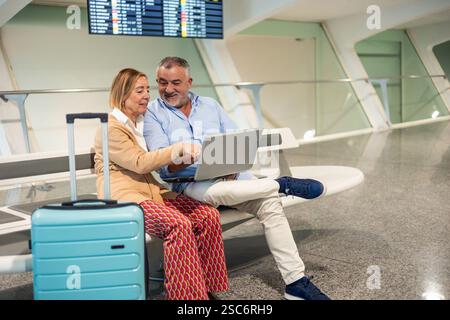 Seniorenpaar sitzt auf der Bank mit Laptop und überprüft Fluginformationen auf dem Bildschirm, mit ihrem Trolley-Koffer daneben, am Flughafen Stockfoto
