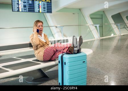 Entspannte Seniorin liegt auf einer Flughafenbank mit den Beinen auf einem Koffer, spricht am Telefon, während sie auf ihren Flug wartet Stockfoto
