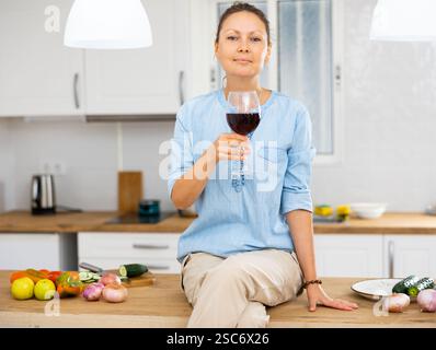 Traumhafte Frau in blauer Bluse, die auf einem Küchentisch sitzt und ein Glas Rotwein genießt Stockfoto