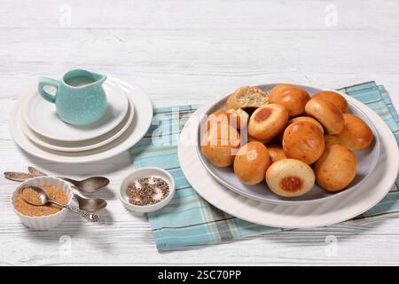 Süße Kardamombrötchen, Pulla, auf einem Teller auf weißem Holztisch mit frischer Creme in blauer Kanne, Serviette, braunem Zucker und Löffeln, horizontale Ansicht von oben, Stockfoto