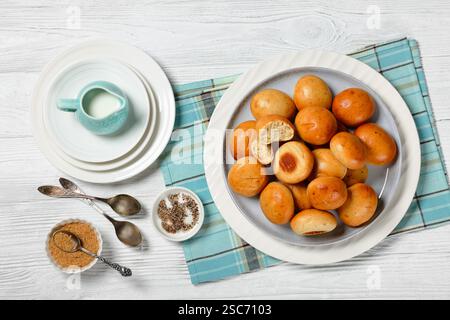 Süße Kardamombrötchen, Pulla, auf einem Teller auf weißem Holztisch mit frischer Creme in blauer Kanne, Serviette, braunem Zucker und Löffeln, horizontale Ansicht von oben, Stockfoto