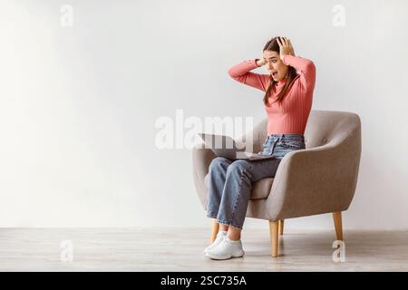 Porträt einer schockierten kaukasischen Frau, die im Sessel mit Laptop sitzt und den Kopf gegen die weiße Studiowand packt Stockfoto