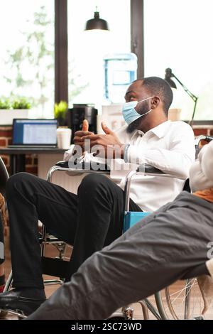 Selektiver Fokus des schwarzen Mannes im Rollstuhl, der über psychische Gesundheit beim Beratungsprogramm in hell beleuchtetem Raum spricht. Porträt eines afroamerikanischen Patienten mit Gesichtsmaske, der seine Probleme bei der Therapie teilt. Stockfoto
