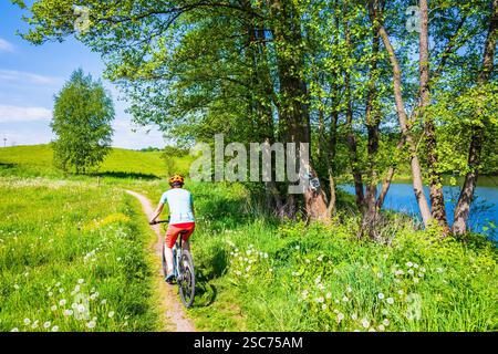 Radfahrerin auf dem Weg entlang des Sees und zwischen grünen Wiesen, Suwalski Landscape Park, Podlasie, Polen Stockfoto