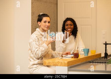 Zwei weibliche Mitbewohnerinnen sitzen an der Küchentheke, genießen Frühstück und führen in ihrem hellen und modernen Apartment, ra, ein lebhaftes Gespräch Stockfoto