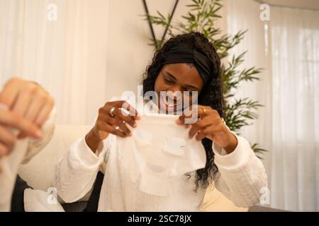 Lächelnde junge schwarze Frau mit weißem Bademantel und Haarreif, die eine kosmetische Gesichtsmaske hält, während einer Schönheitsbehandlung zu Hause auf einem Sofa sitzt Stockfoto