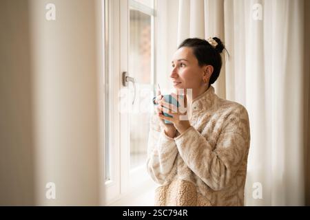 Eine junge Frau mit beigefarbenem Pullover genießt an einem Wintermorgen eine Tasse heißes Getränk, während sie aus dem Fenster in ihrem gemütlichen Zuhause schaut und sich entspannt fühlt Stockfoto