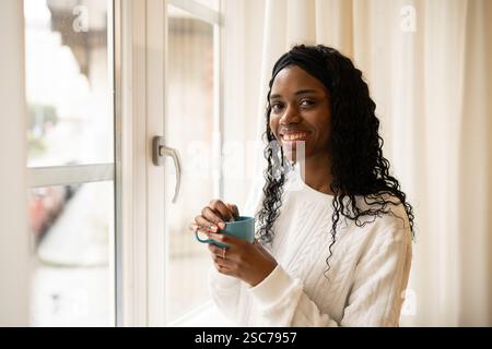 Eine junge Frau mit langen, lockigen Haaren und einem stilvollen Stirnband, die einen Becher hält und ein heißes Getränk genießt, während sie einen ruhigen Moment am Fenster in sich hat Stockfoto