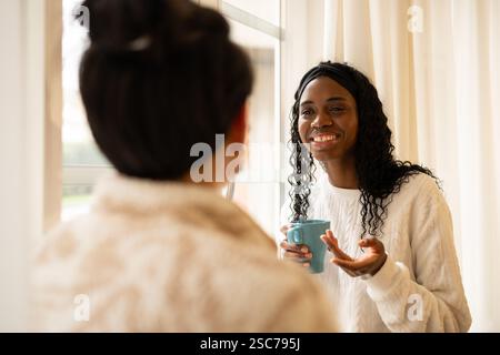Zwei Frauen, die in bequemen Pullover gekleidet sind, führen eine freundliche Unterhaltung in der Nähe eines Fensters, eine hält eine Kaffeetasse und teilt einen Moment der Verbindung Stockfoto