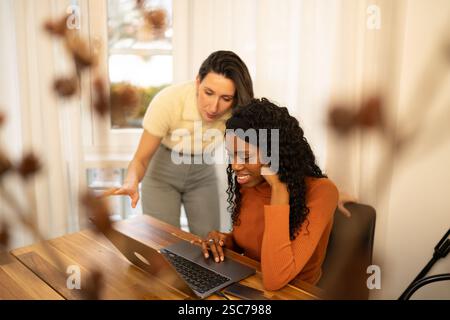 Zwei weibliche Kollegen arbeiten an einem Projekt mit einem Laptop in einem Home Office zusammen, einer zeigt auf den Bildschirm, während der andere aufmerksam beobachtet, hoch Stockfoto