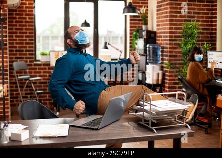 Junger männlicher Unternehmer steht im gemauerten Raum und feiert erfolgreiche Präsentation. Geschäftsmann trägt Gesichtsmaske und tanzt im Startup-Büro, nachdem er ein Geschäftsprojekt gewonnen hat. Stockfoto