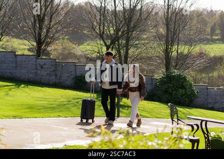 Junge Paare gehen Hand in Hand durch den Garten eines Hotels, ziehen einen Koffer und genießen die herbstliche Atmosphäre während ihres Urlaubs Stockfoto