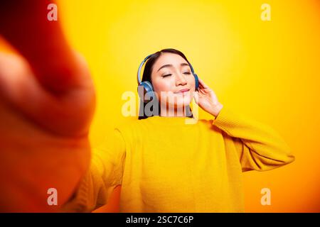 Junge Frau, die mit geschlossenen Augen Musik hört und täglich einen Vlog für ihre Zuschauer macht. Asiatische Dame mit Kopfhörern und Kamera für ein Selfie vor gelbem Hintergrund. Stockfoto