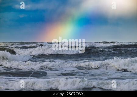 Ein lebhafter Regenbogen überzieht einen turbulenten Ozean mit stürzenden Wellen unter einem bewölkten Himmel. Stockfoto
