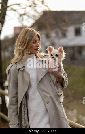 Eine junge Frau in einem stilvollen Trenchcoat hält einen Chihuahua, während sie durch einen friedlichen Park mit Bäumen und einem Haus im Hintergrund auf einem Su spaziert Stockfoto