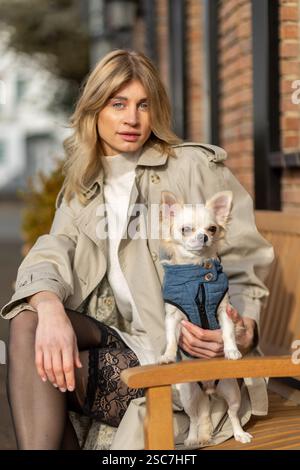 Eine Frau mit stilvollem Trenchcoat und gemusterter Strumpfhose sitzt auf einer Bank und hält einen kleinen Chihuahua in einer blauen Weste. Der Hintergrund zeigt urb Stockfoto