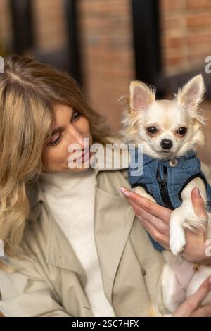 Eine Frau in einer leichten Jacke sitzt draußen und hält einen kleinen Hund in einem Jeansmantel. Sie lächelt liebevoll und genießt den Moment in A Stockfoto