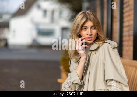 Eine junge Frau mit langen blonden Haaren sitzt auf einer Bank und trägt einen beigefarbenen Trenchcoat. Sie führt ein Telefongespräch, während sie einen sonnigen Tag in sich verbringt Stockfoto