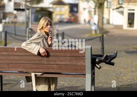 Eine junge Frau entspannt sich auf einer Holzbank in einem Stadtpark in stilvoller Kleidung. Sie benutzt gern ein Vape-Gerät, während sie ihre Umgebung im beobachtet Stockfoto