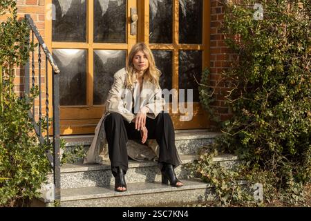 Eine Frau sitzt lässig auf den Stufen eines charmanten Backsteinhauses, trägt einen modischen Trenchcoat und eine schwarze Hose. Das Sonnenlicht hebt sie entspannt hervor Stockfoto