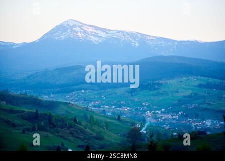 Szene des Taldorfes, umgeben von grünen Hügeln, mit majestätischen, schneebedeckten Bergen im Hintergrund. Das Licht am frühen Morgen oder am späten Abend strahlt ein heiteres Leuchten aus, während Rauchschauer aus den Schornsteinen steigen. Stockfoto
