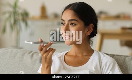 Glückliche lächelnde indische Frau zu Hause Arabische positive arabische Mädchen weibliche Dame im Wohnzimmer Handy sprechendes Mikrofon Smartphone Mikrofon Mikrofon sprechen Stockfoto