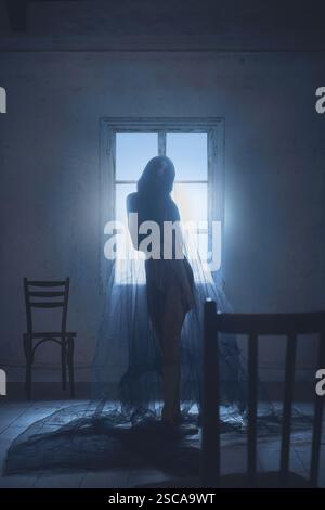 Silhouette einer Frau mit langen dunklen Haaren, bedeckt mit transparentem Schleier, stehend in leerem Raum mit zwei Sesseln und Fenster. Stockfoto