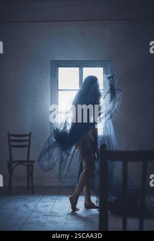Silhouette einer Frau mit langen dunklen Haaren, bedeckt mit transparentem Schleier, stehend in leerem Raum mit zwei Sesseln und Fenster. Stockfoto