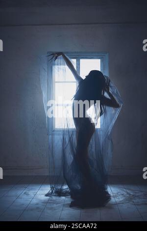 Silhouette einer Frau mit langen dunklen Haaren, bedeckt mit transparentem Schleier, stehend in leerem Raum mit zwei Sesseln und Fenster. Stockfoto