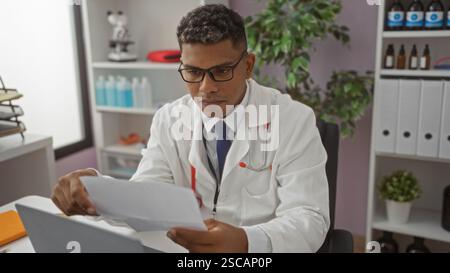 Junger hispanischer Mann Doktor arbeitet drinnen in einer Klinik, trägt einen weißen Mantel und eine Brille, überprüft Papiere in einer medizinischen Praxis mit verschiedenen medizinischen Sup Stockfoto