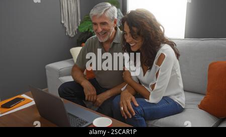 Paare mittleren Alters genießen die Zeit drinnen zusammen und beobachten den Laptop lächelnd in einem gemütlichen Wohnzimmer mit modernem Dekor und warmem Ambiente Stockfoto