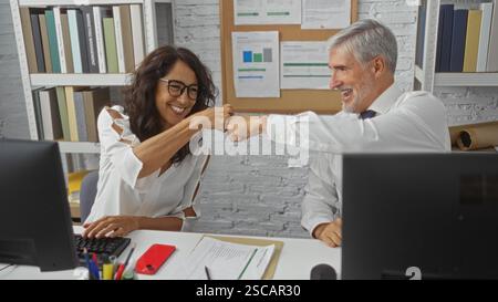 Frau und Mann lächeln und Faustschläge in einem modernen Büroumfeld, umgeben von Computern und Papierkram, zeigen eine kollaborative und professionelle Person Stockfoto