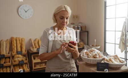 Frau in Bäckerei mit kurzen blonden Haaren benutzt ihr Telefon, während sie von frischem Brot und Gebäck umgeben ist Stockfoto