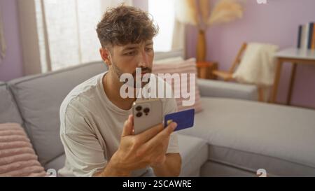 Junger Mann, der in einem Wohnzimmer sitzt und ein Smartphone und eine Kreditkarte mit ernsthaftem Ausdruck hält Stockfoto