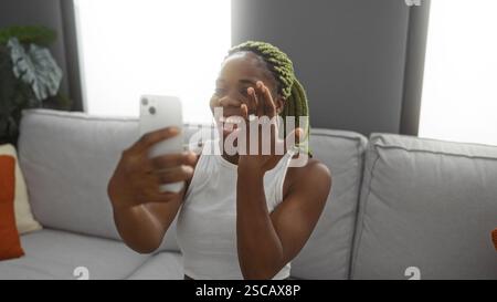 Frau, die während eines Videoanrufs mit dem Smartphone einen Verlobungsring zeigt, fröhlich lächelt, bequem in einem hellen, modernen Wohnzimmer sitzt, aufgeregt, glücklich Stockfoto