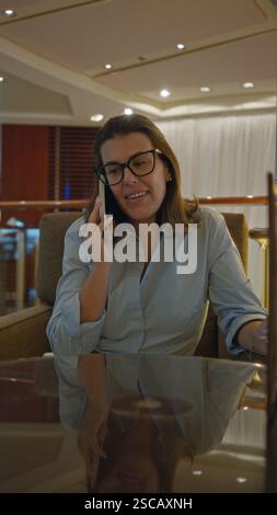Frau telefoniert in einer luxuriösen Bar, lächelt und entspannt, trägt Brille und legere Kleidung, sitzt drinnen und spiegelt moderne Eleganz wider. Stockfoto