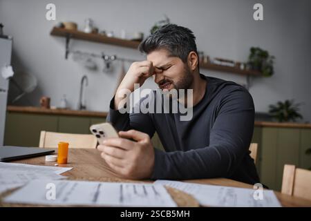 Ein gutaussehender Mann sitzt an einem Holztisch, gestresst, während er Dokumente liest und sein Handy überprüft. Stockfoto