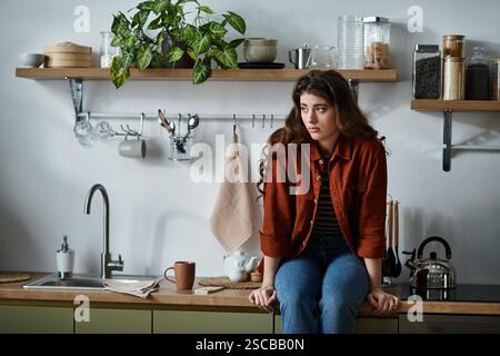 Eine junge Frau sitzt auf dem Theken in einer sanft beleuchteten Küche, verloren in Gedanken in einer ruhigen Einrichtung. Stockfoto