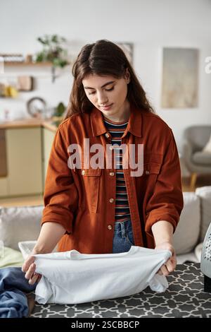 Eine junge Frau verarbeitet ihre Emotionen, während sie in ihrem einladenden Wohnzimmer ein Hemd faltet. Stockfoto