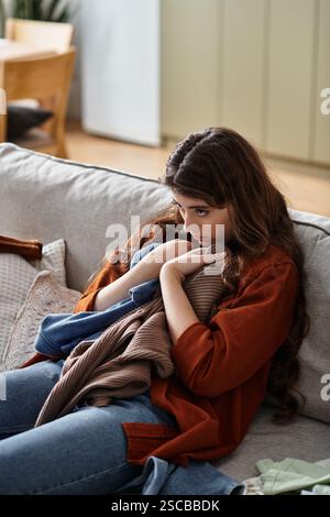 Eine Frau sitzt leise auf einem Sofa und nimmt Kleidung an, während sie über ihre Traurigkeit reflektiert. Stockfoto