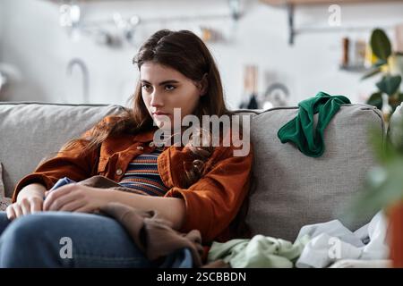 Eine Frau sitzt still auf einer Couch, umgeben von unordentlichen Kleidern, und zeigt ihre innere Traurigkeit. Stockfoto