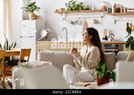Eine Frau sitzt leise auf einer Couch und denkt tief nach, während sie ein warmes Getränk hält. Stockfoto