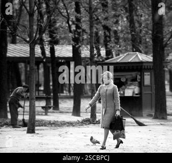 Frau mit Mantel und Taschen, die in Paris durch den Jardin du Luxembourg geht. Hinten eine Kehrmaschine. Stockfoto